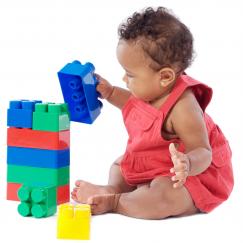 On a blank white background a baby girl sits and places a blue plastic brick upon a stack of other bricks of blue, green and red. Nearby a yellow block sits on the floor.